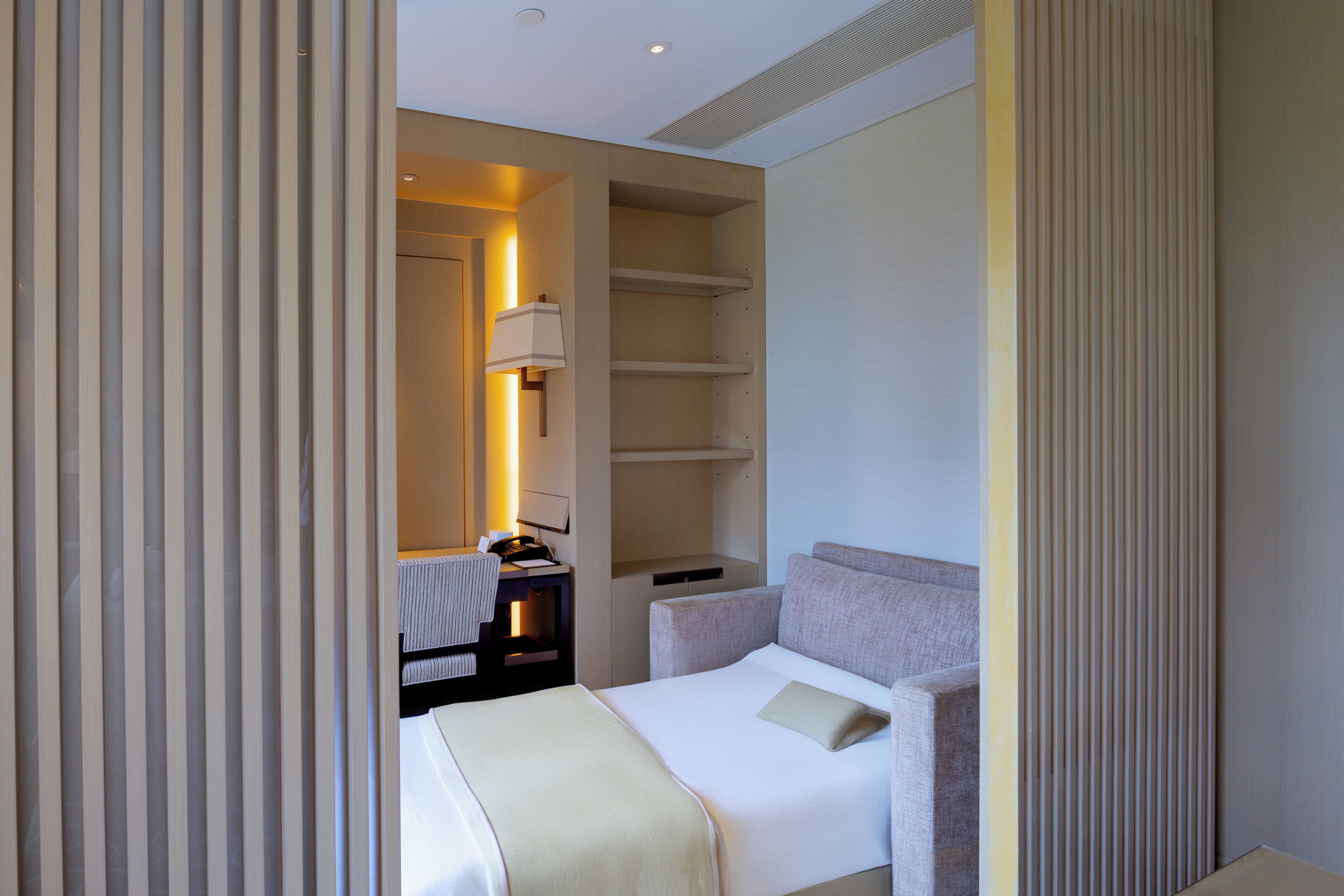 Une chambre d’hôtel moderne avec un lit soigneusement fait, une table de chevet avec une lampe et une bibliothèque intégrée.