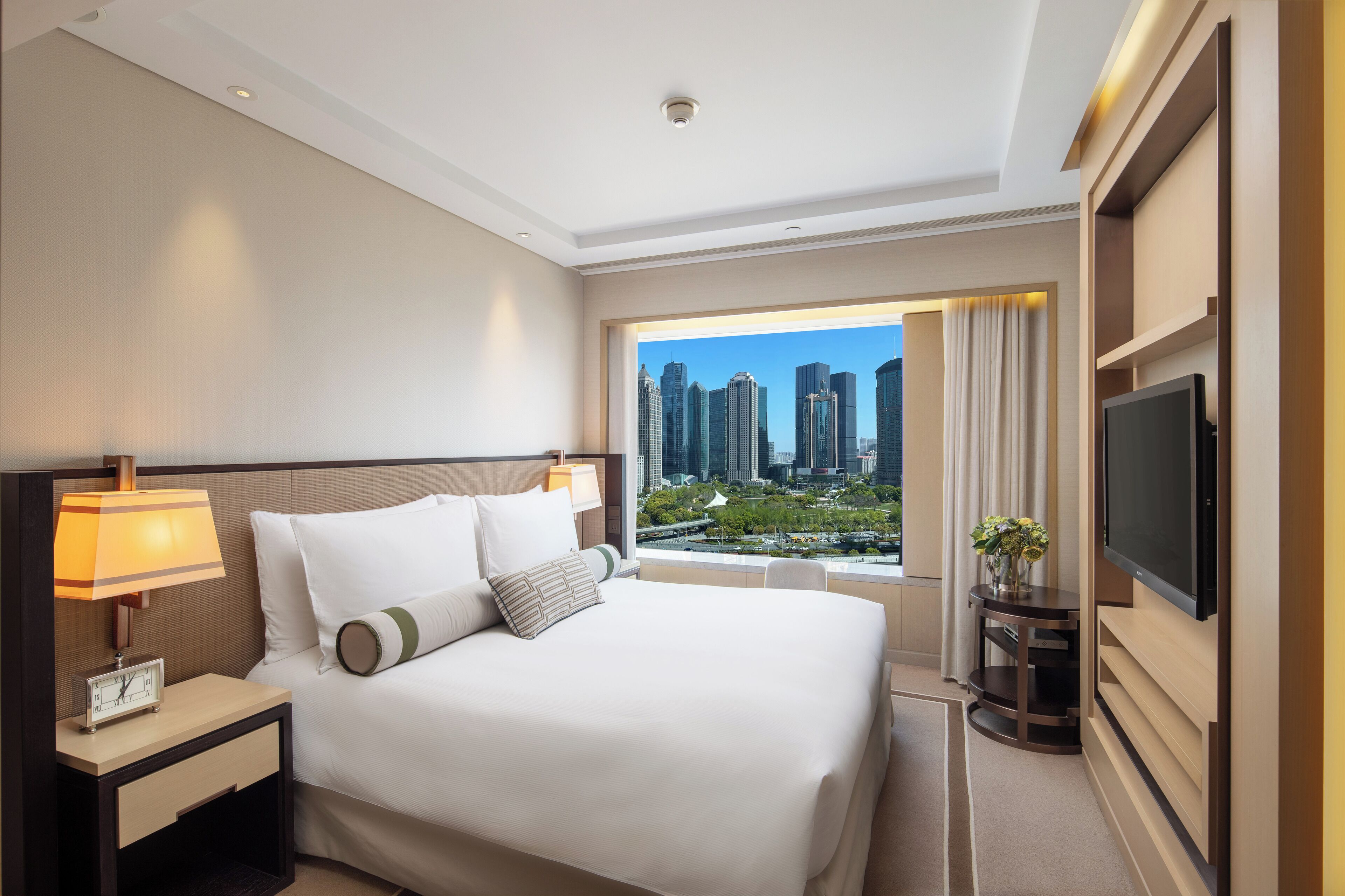 Habitación de hotel con una cama grande, televisión, mesita de noche con lámpara y vistas a la ciudad.