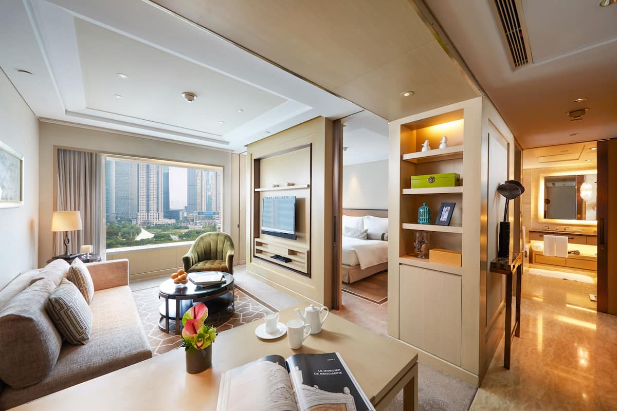 Una habitación de hotel moderna con un ventanal grande, un sofá, una mesita de centro, un rincón de lectura, un televisor y una kitchenette.