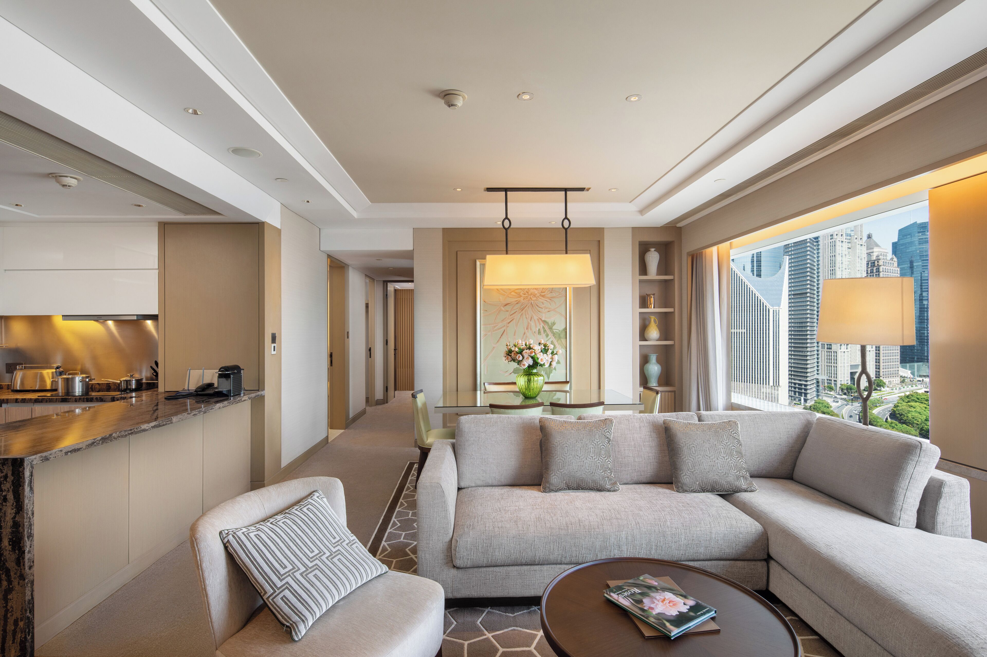 A modern hotel room with a living area, a kitchen, and a large window with a city view.
