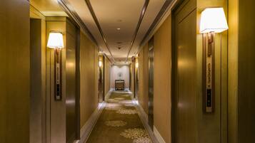 A hotel corridor with elevator doors on both sides, a carpeted floor, and wall-mounted lamps.