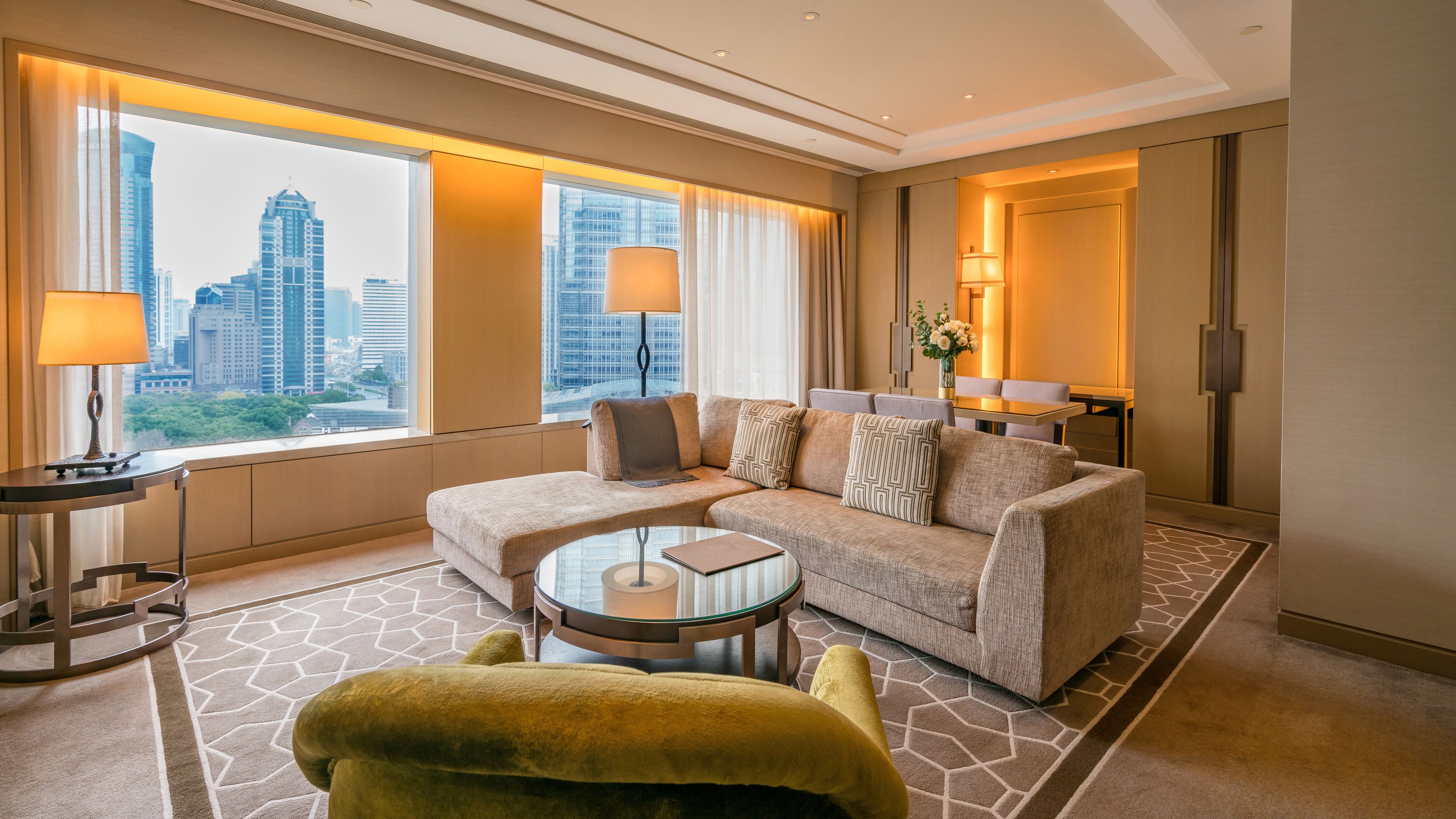 A modern hotel room with a large window offering a city view, a sofa, a glass coffee table, and a small round table with a lamp.