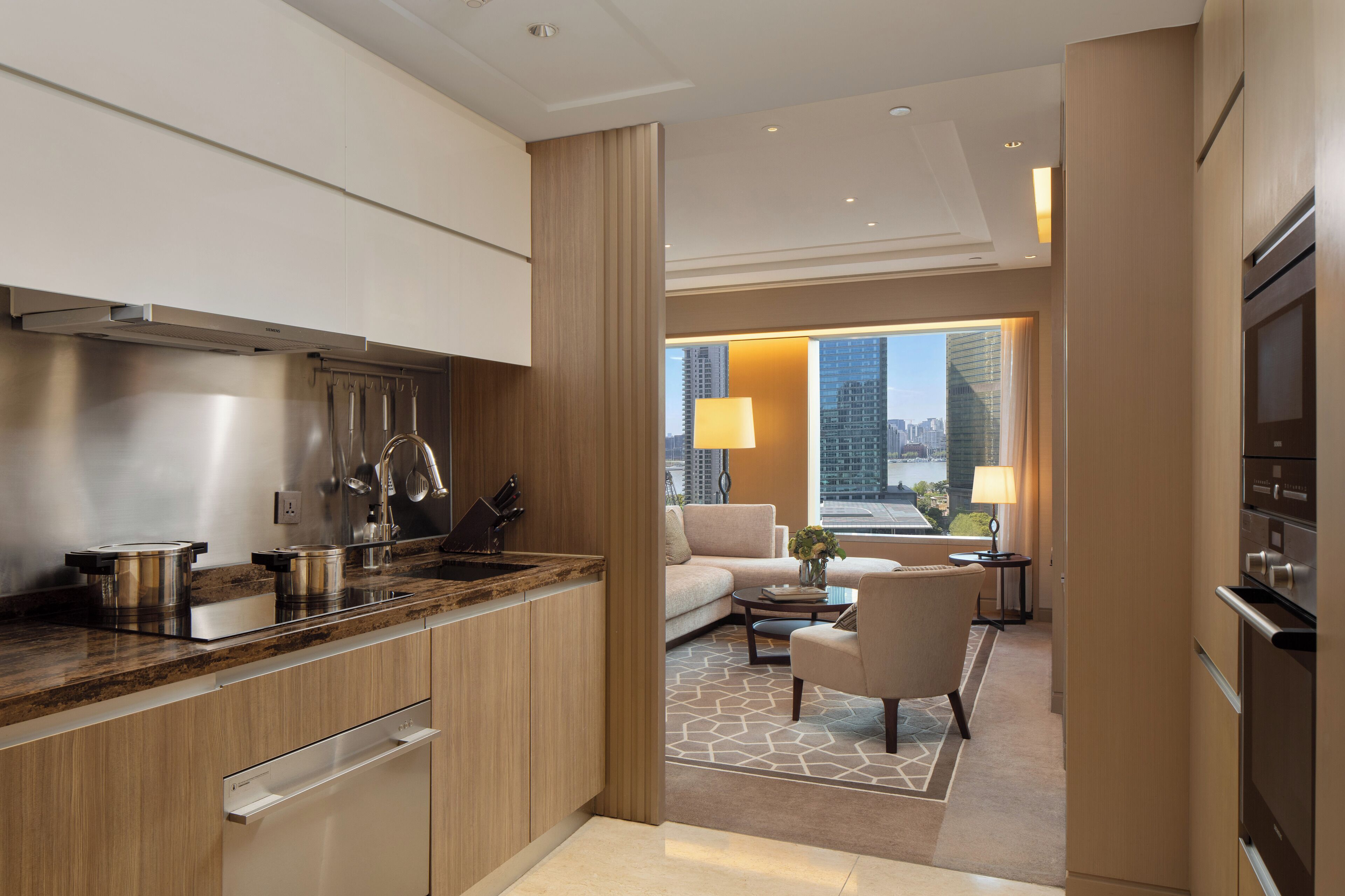 Une chambre d’hôtel moderne dotée d’une cuisine, d’un espace de vie avec un canapé et des fauteuils, et d’une grande fenêtre donnant sur la ville.