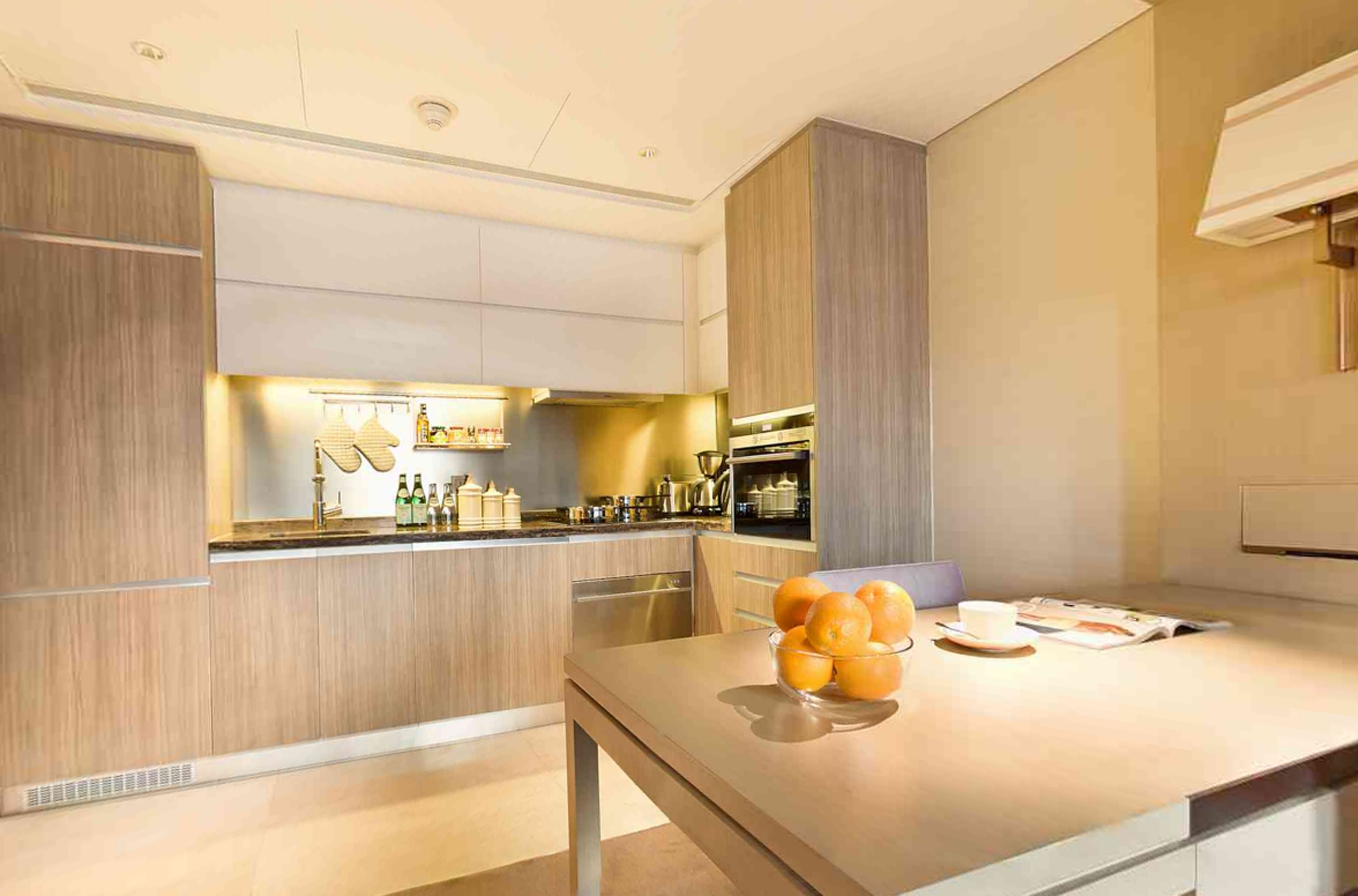 A modern kitchen with built-in appliances, a central island with a bowl of fruit, and a bar area with bottles.