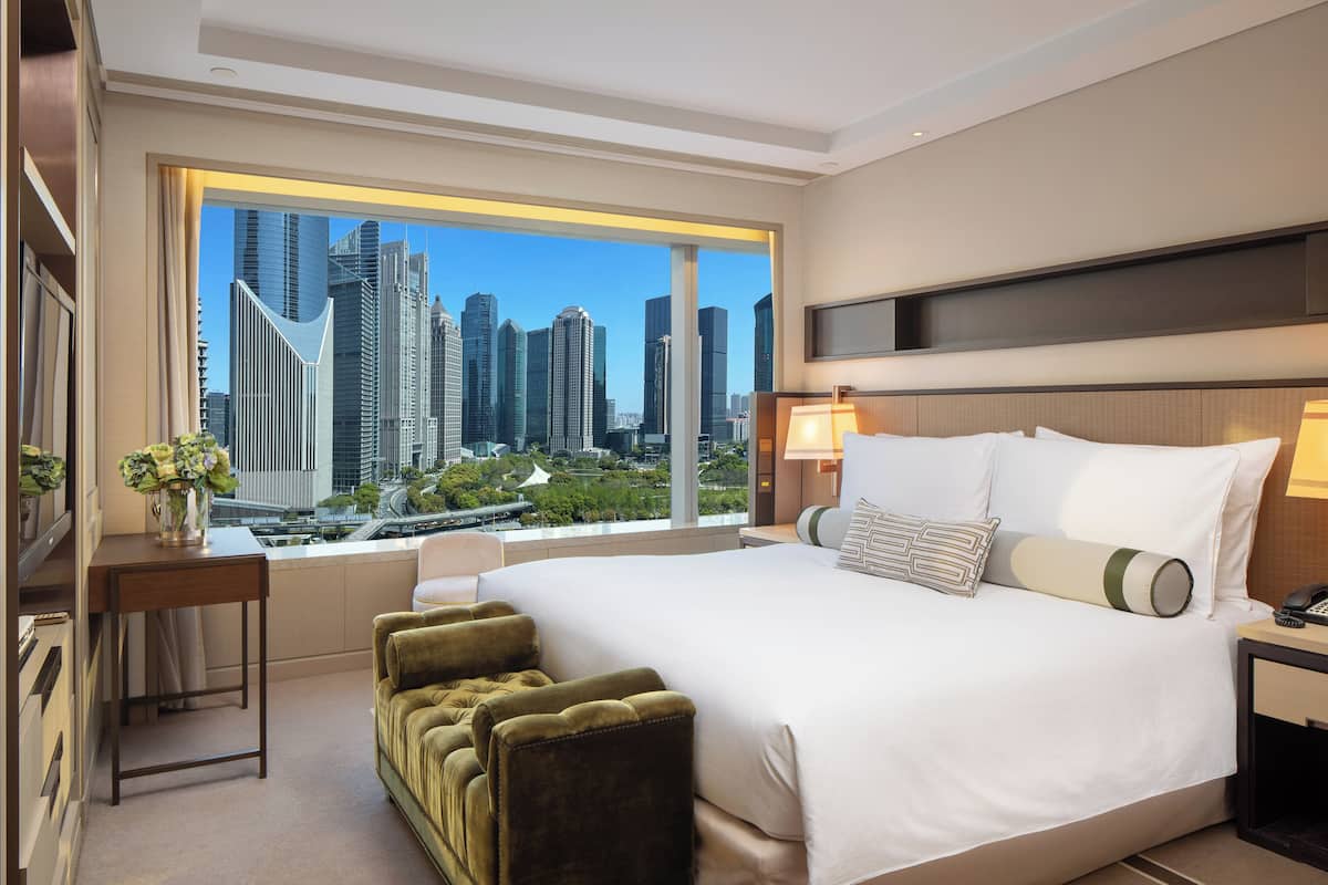 Una habitación de hotel moderna con una cama grande, vistas a un paisaje urbano, un escritorio con un florero y un banco tapizado en verde.