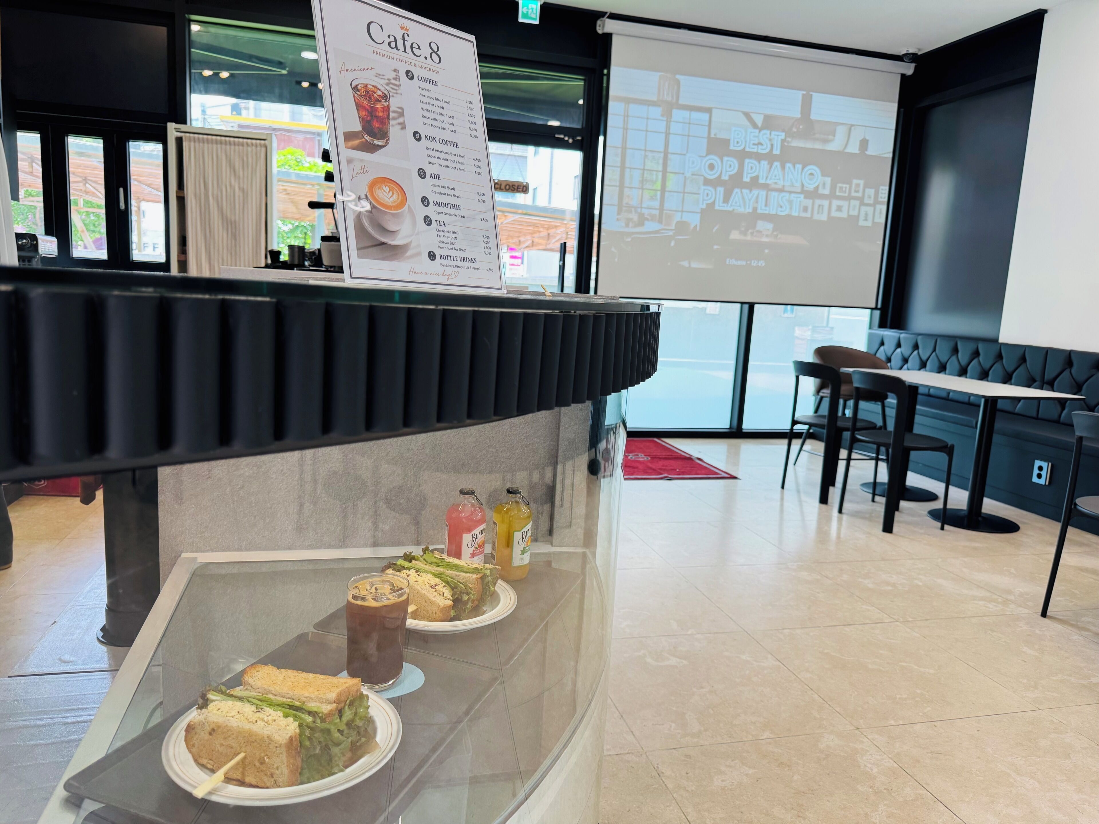 Een café met een menubord, een scherm waarop een afspeellijst staat en een toonbank met belegde broodjes en drankjes.