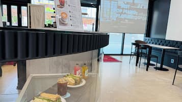 Een café met een menubord, een scherm waarop een afspeellijst staat en een toonbank met belegde broodjes en drankjes.