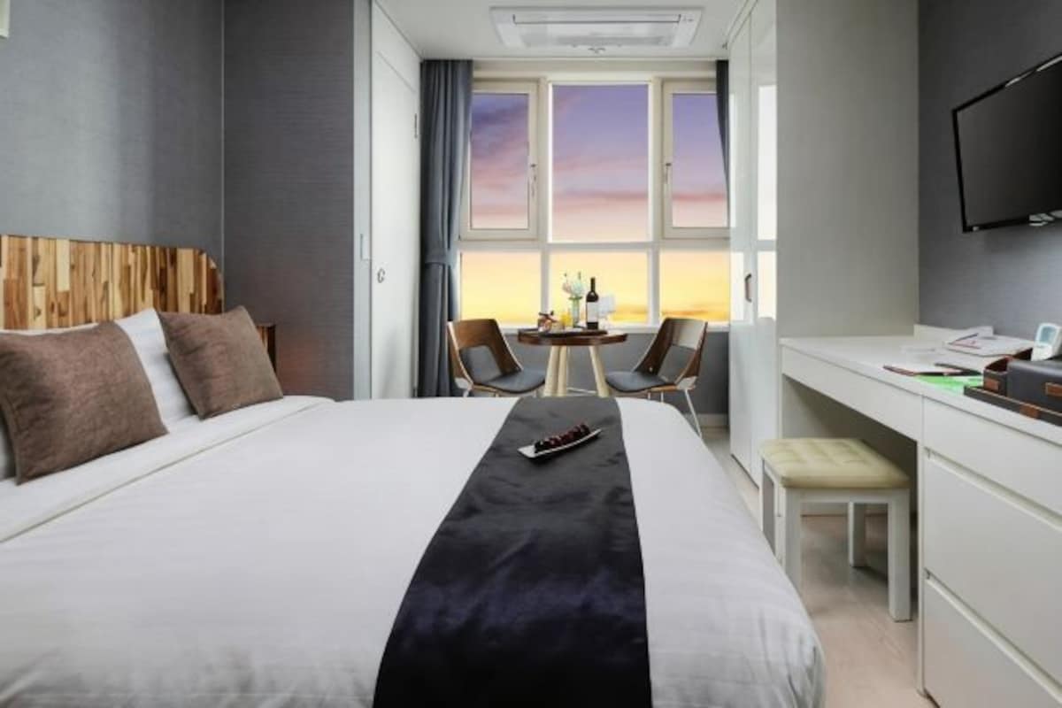 A hotel room with a bed, desk, chairs, and a view of the sunset through the window.
