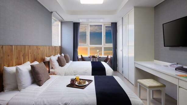 A hotel room with two beds, a desk, a TV, and a view of the sunset through the window. - Shinchon Ever8 Serviced Residence (Seoul)