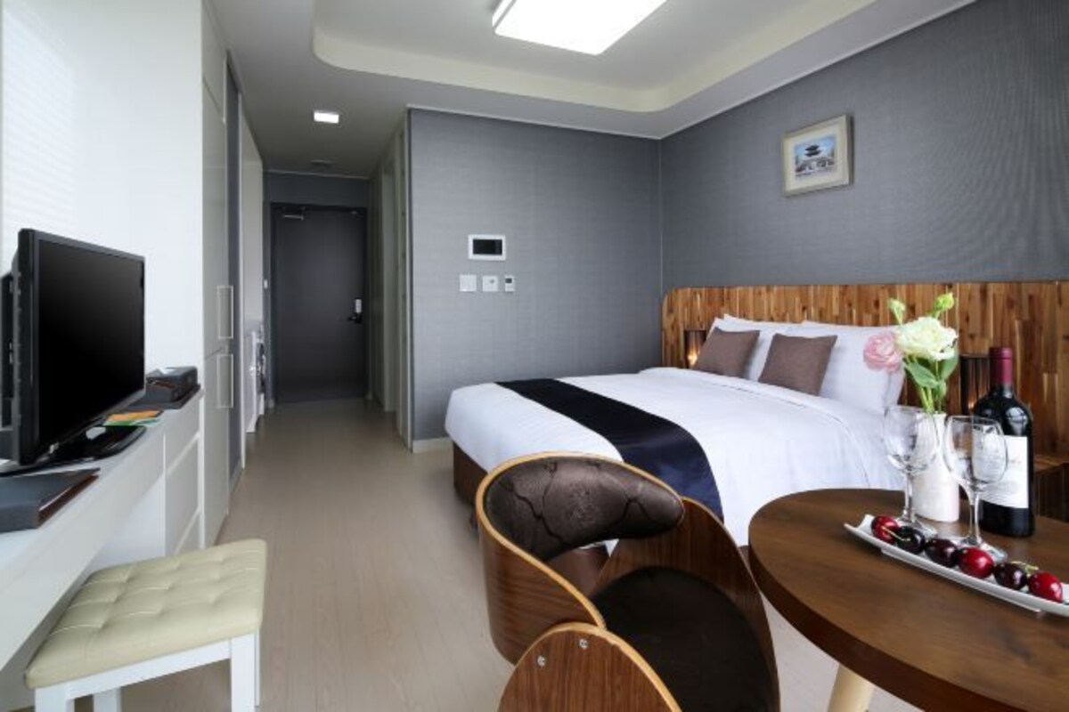 A hotel room with a bed, a television, a desk with a chair, and a dining table with a wine bottle and glasses.