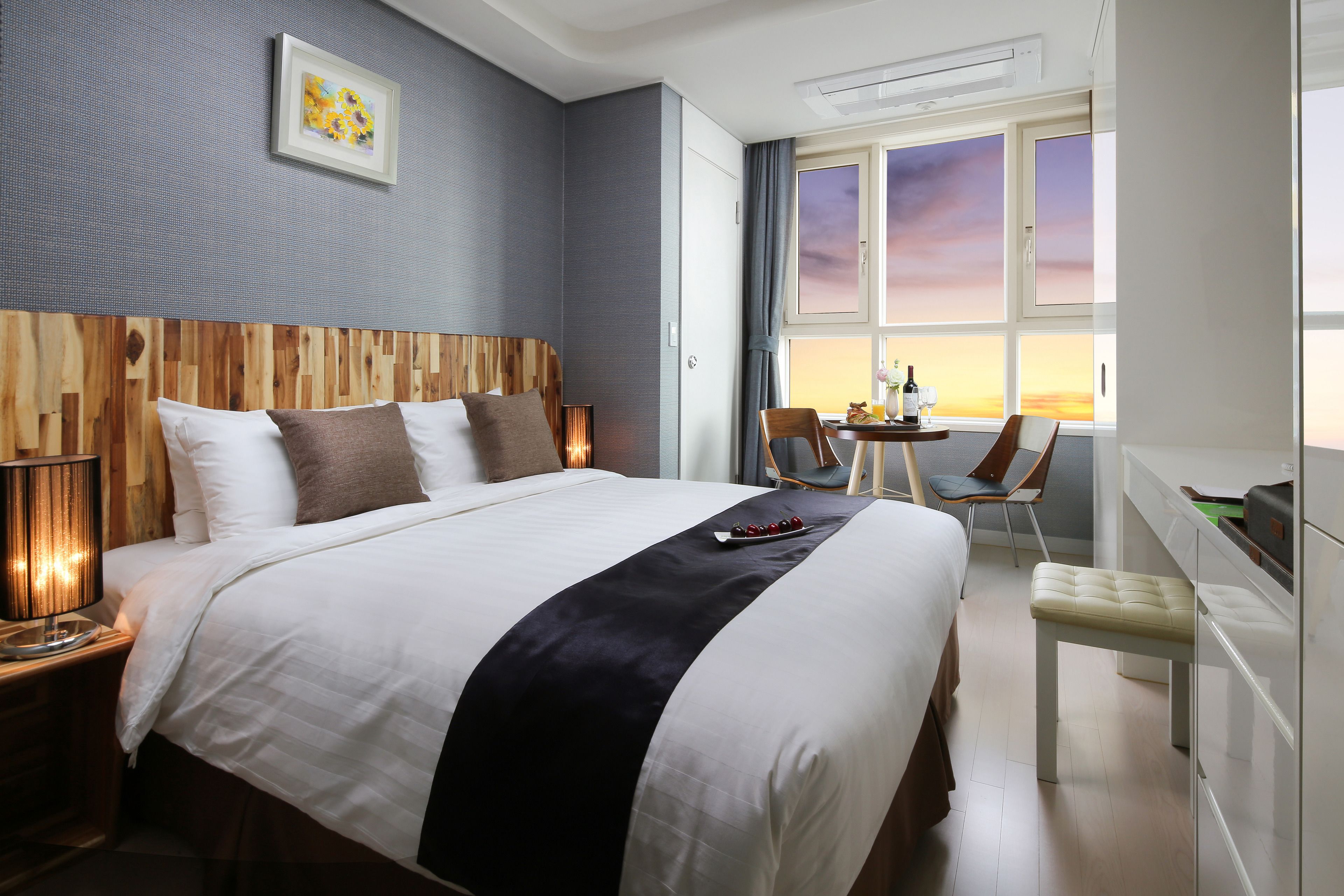 Ein Hotelzimmer mit einem großen Bett, einem Schreibtisch mit Stuhl, einem Fenster mit Sonnenuntergangsblick und einem Wandbild.