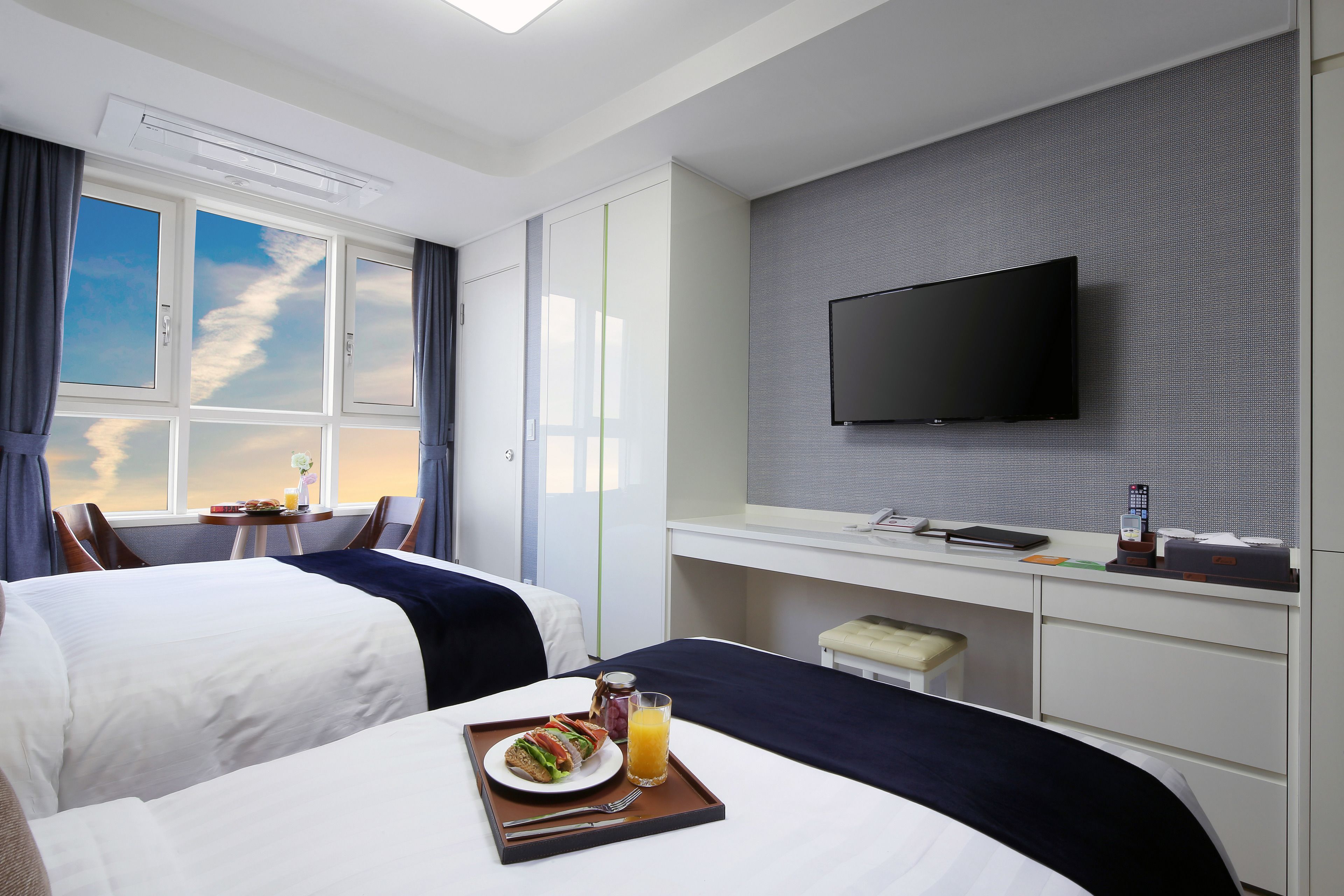 A hotel room with two beds, a desk, a TV, and a breakfast tray with a sandwich and juice.
