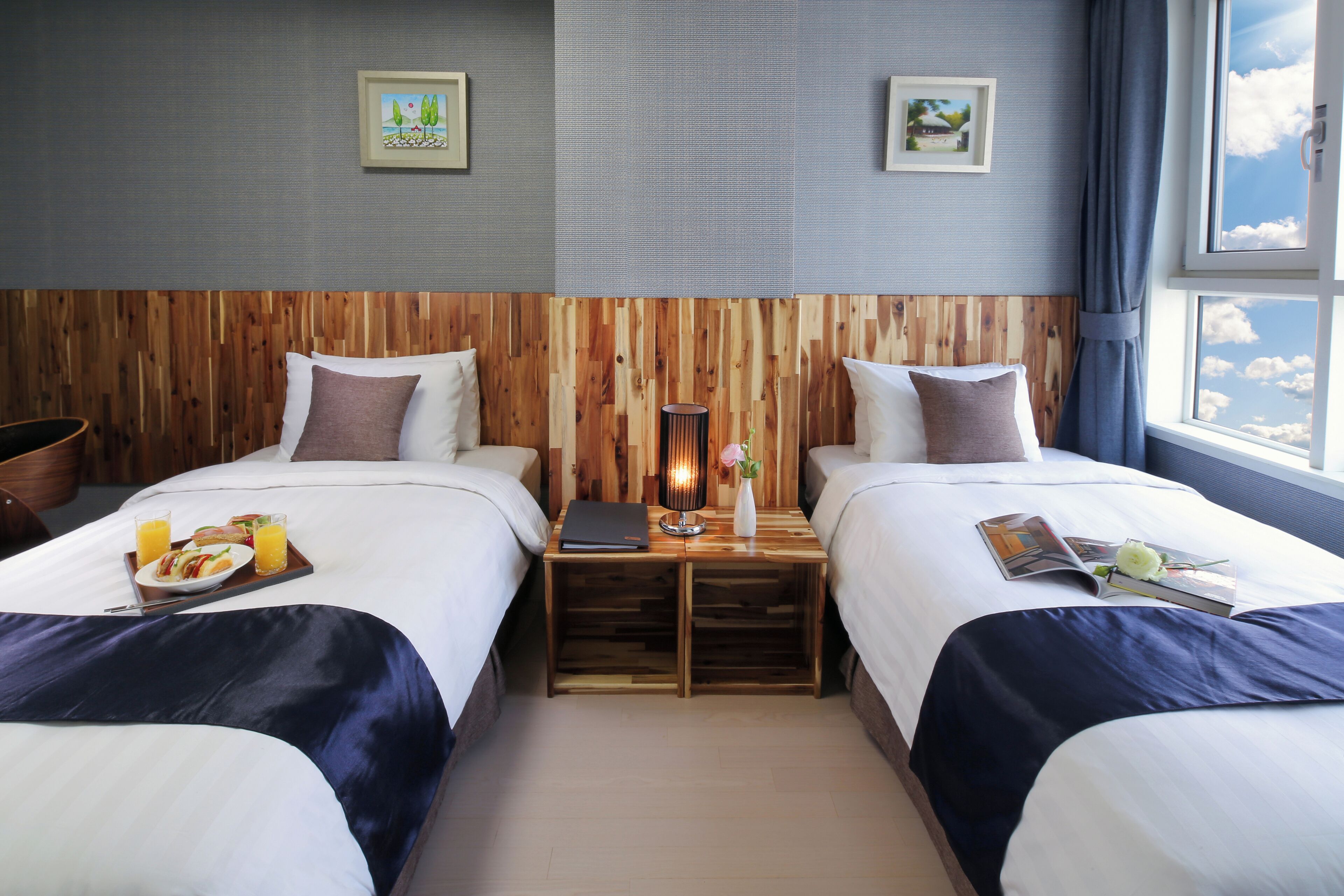 Ein Hotelzimmer mit zwei Betten, einem Holz-Kopfteil, einem kleinen Tisch mit Lampe und einem Fenster mit Blick zum Himmel.