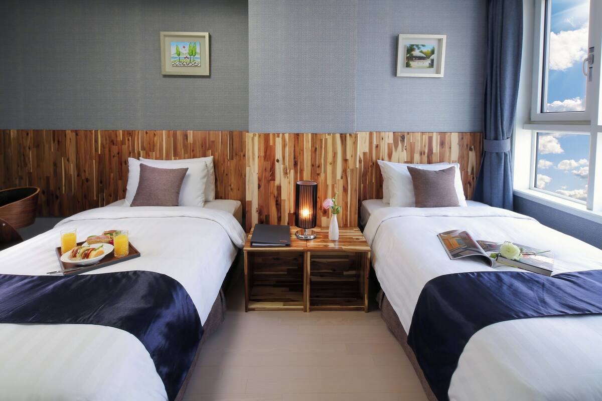A hotel room with two beds, a wooden headboard, a small table with a lamp, and a window with a view of the sky.