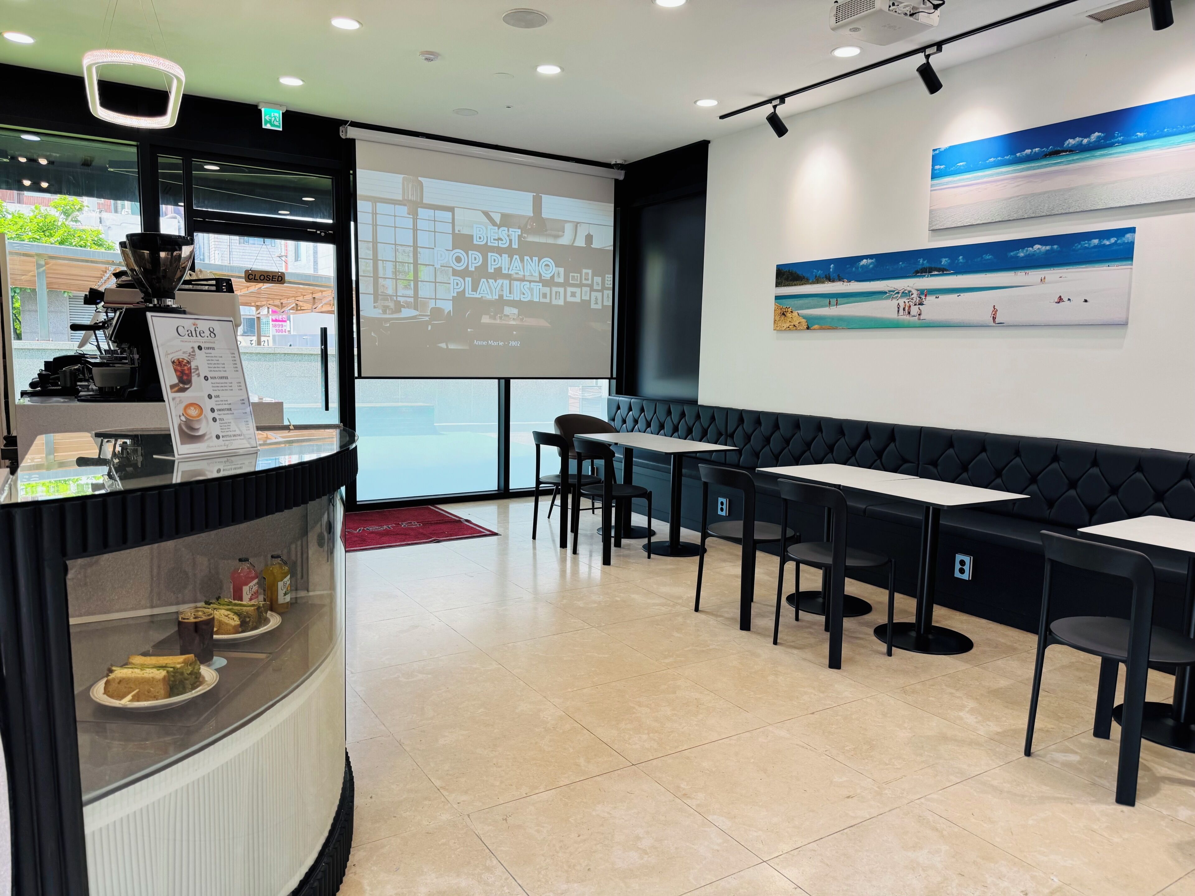 A café with a counter displaying menu items, a projection screen with a presentation, and a seating area with tables and chairs.