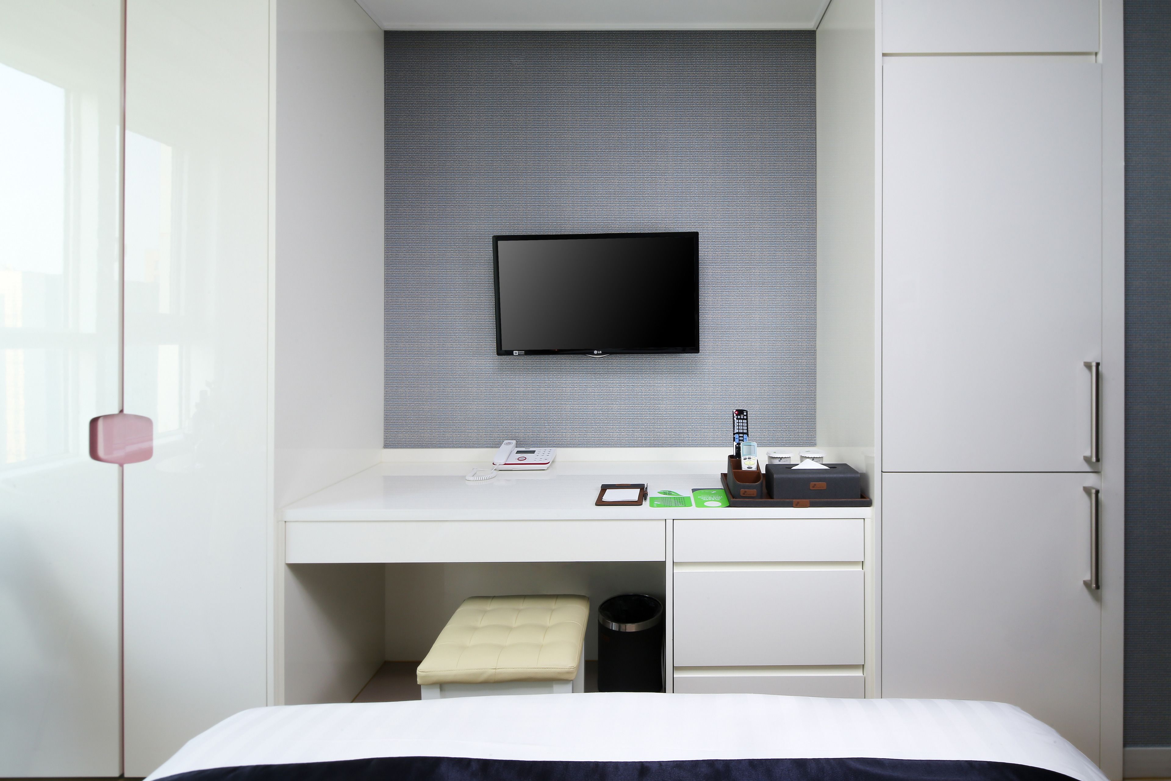A hotel room with a bed, a desk with a flat-screen TV, a small trash bin, and a cabinet.