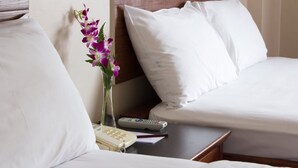 Quarto de hotel com uma cama, mesa de cabeceira com telefone e controle remoto, e um vaso com flores roxas.