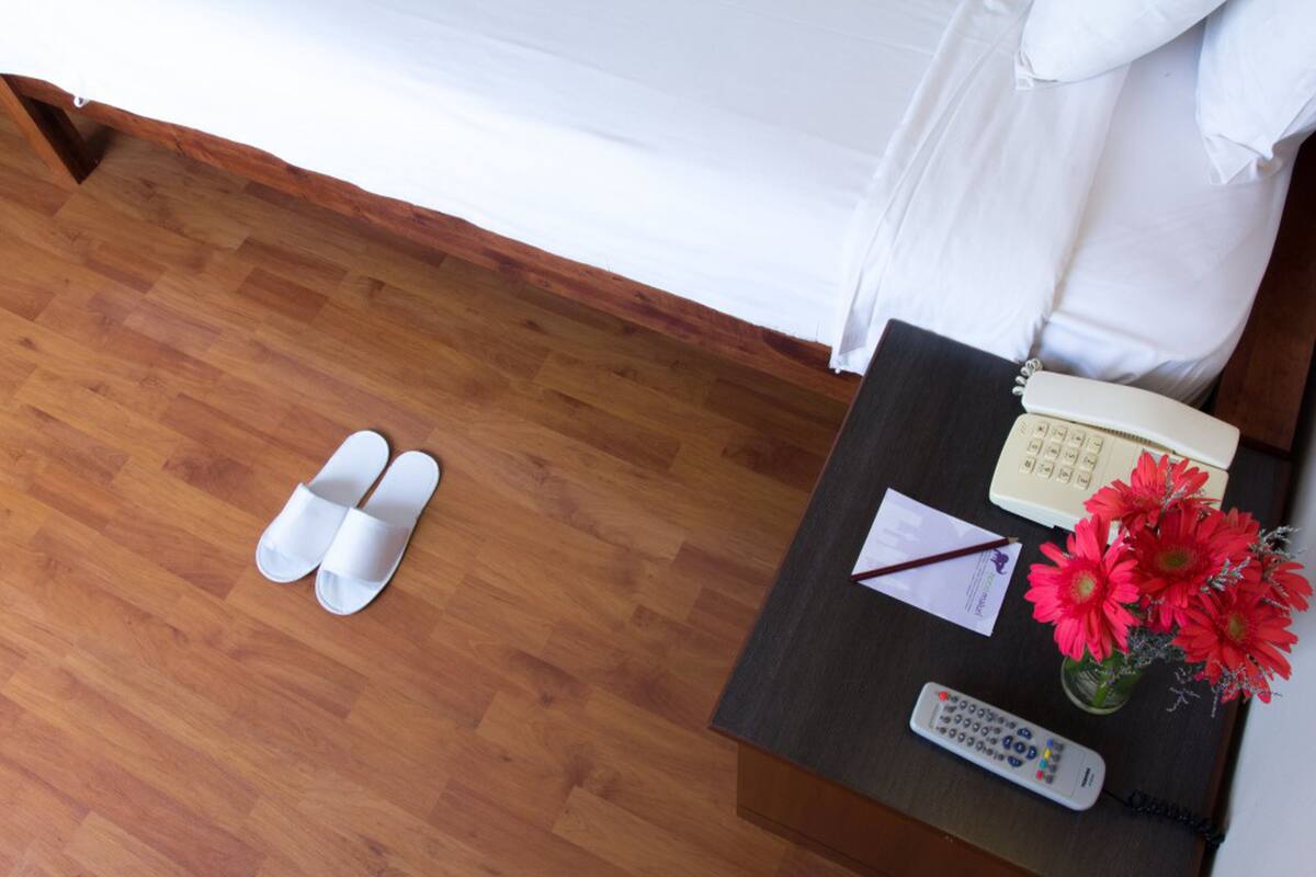 A hotel room with a wooden floor, a bed with white bedding, a nightstand with a telephone, a remote control, a notepad, and a vase with red flowers.