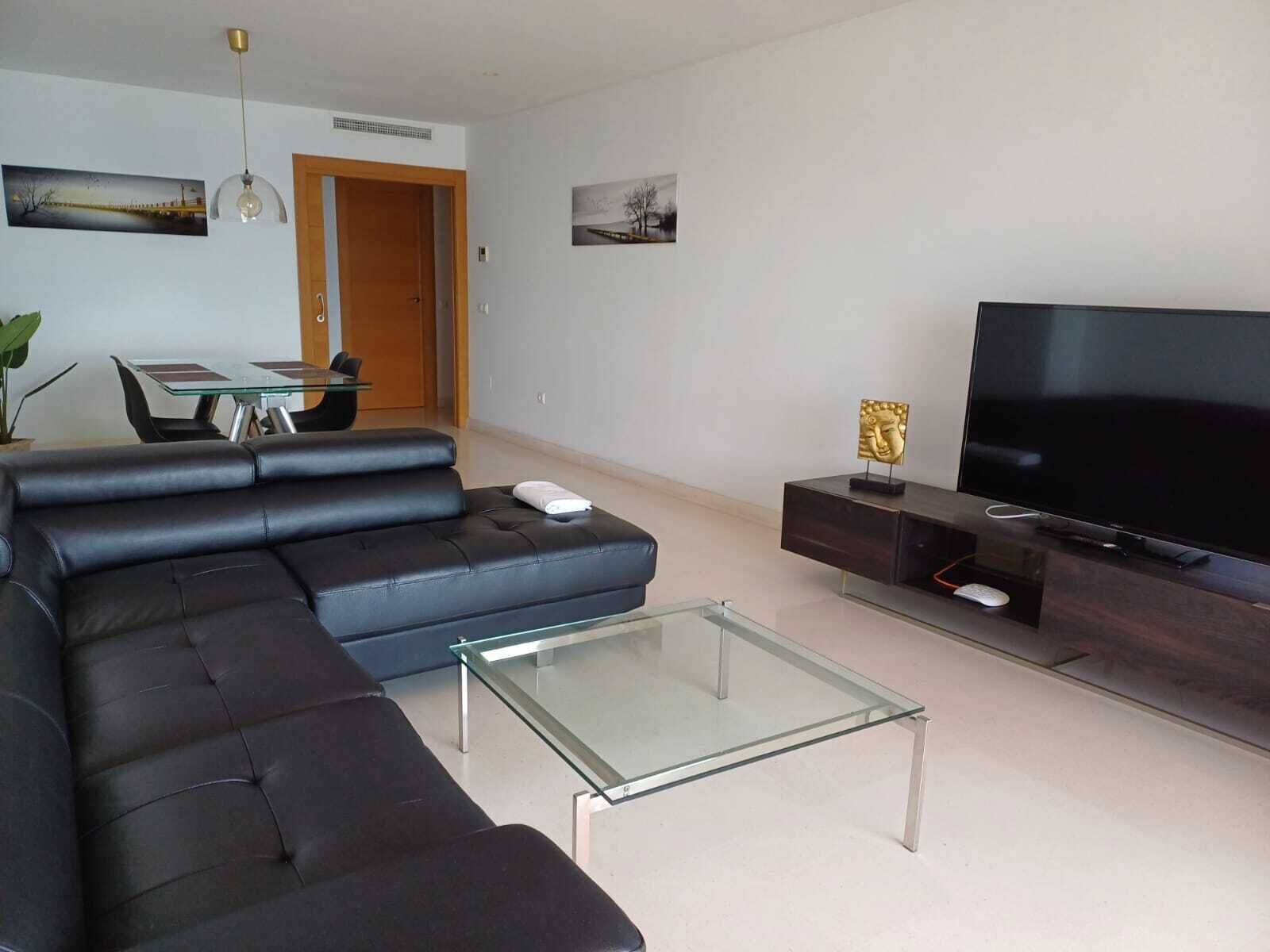 Una sala de estar moderna con un sofá seccional, una mesa de centro de vidrio, un televisor de pantalla plana y un comedor con una mesa y sillas de vidrio.