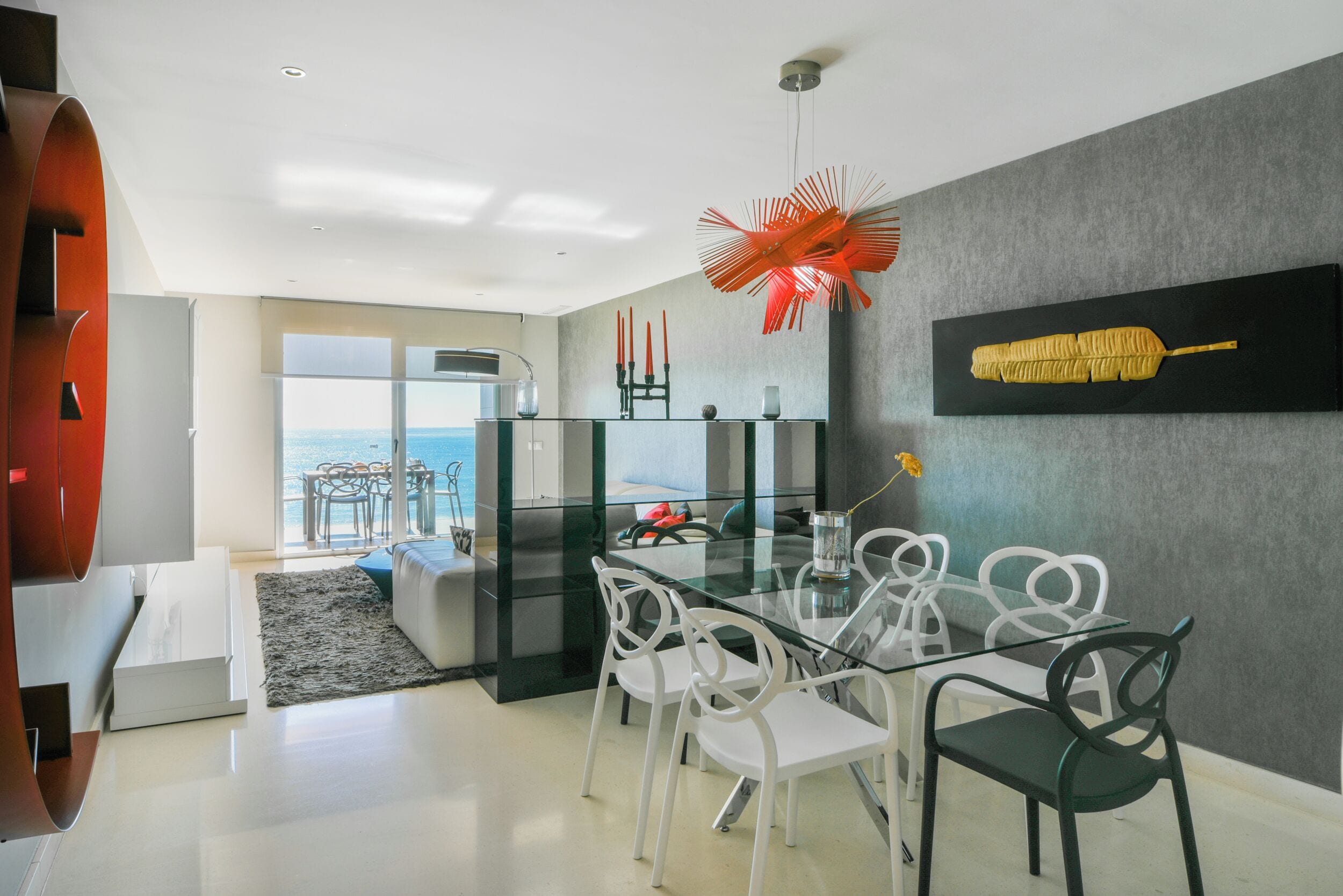 Un comedor moderno con una mesa de vidrio y sillas blancas, una pieza decorativa roja de gran tamaño y una obra de arte enmarcada en la pared.