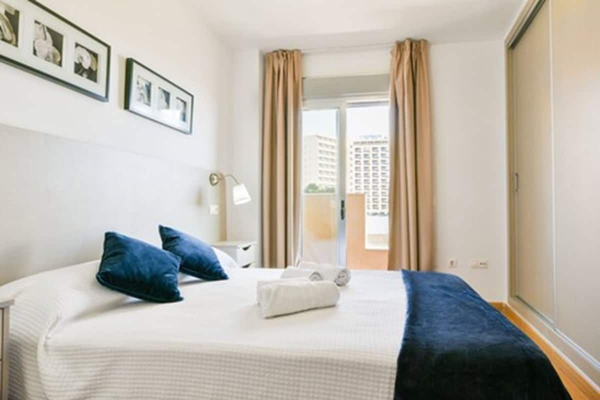 A hotel room with a bed, a bedside table, a lamp, and a view of the outside.