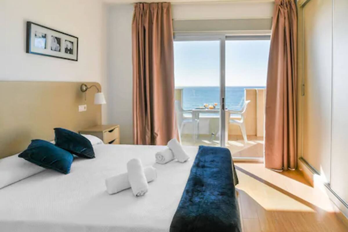 A hotel room with a bed, a bench, a desk, a chair, and a view of the sea.