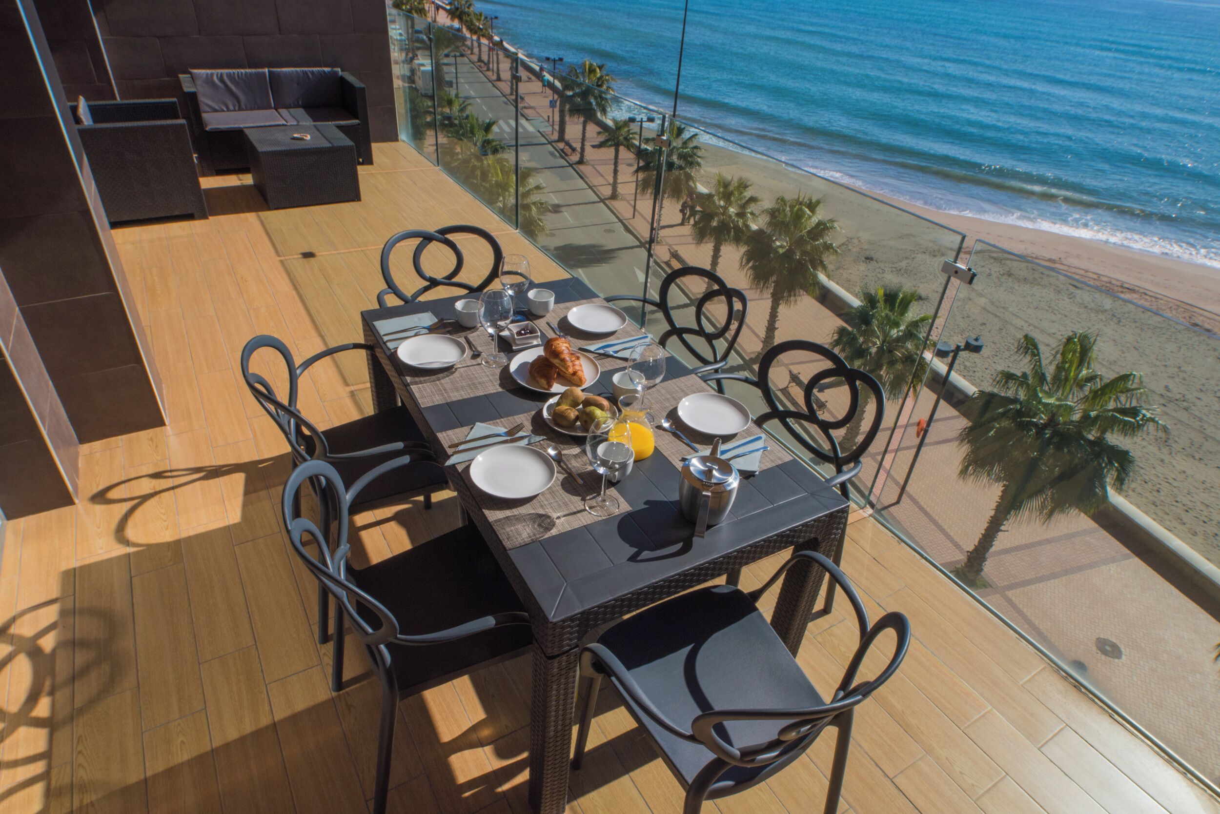 A dining table with chairs on a balcony overlooking a beach.