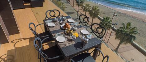 A dining table with chairs on a balcony overlooking a beach.
