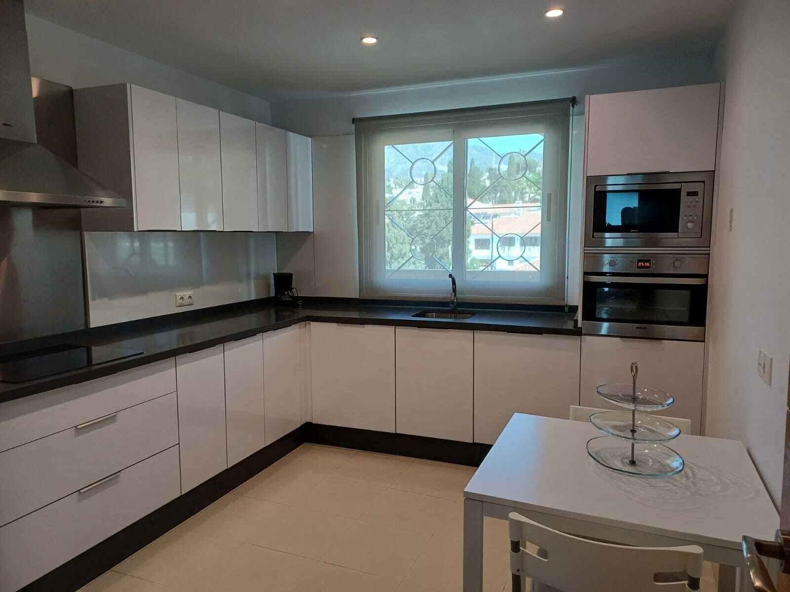A modern kitchen with white cabinets, a black countertop, a window with a view, and a dining table with a glass cake stand.