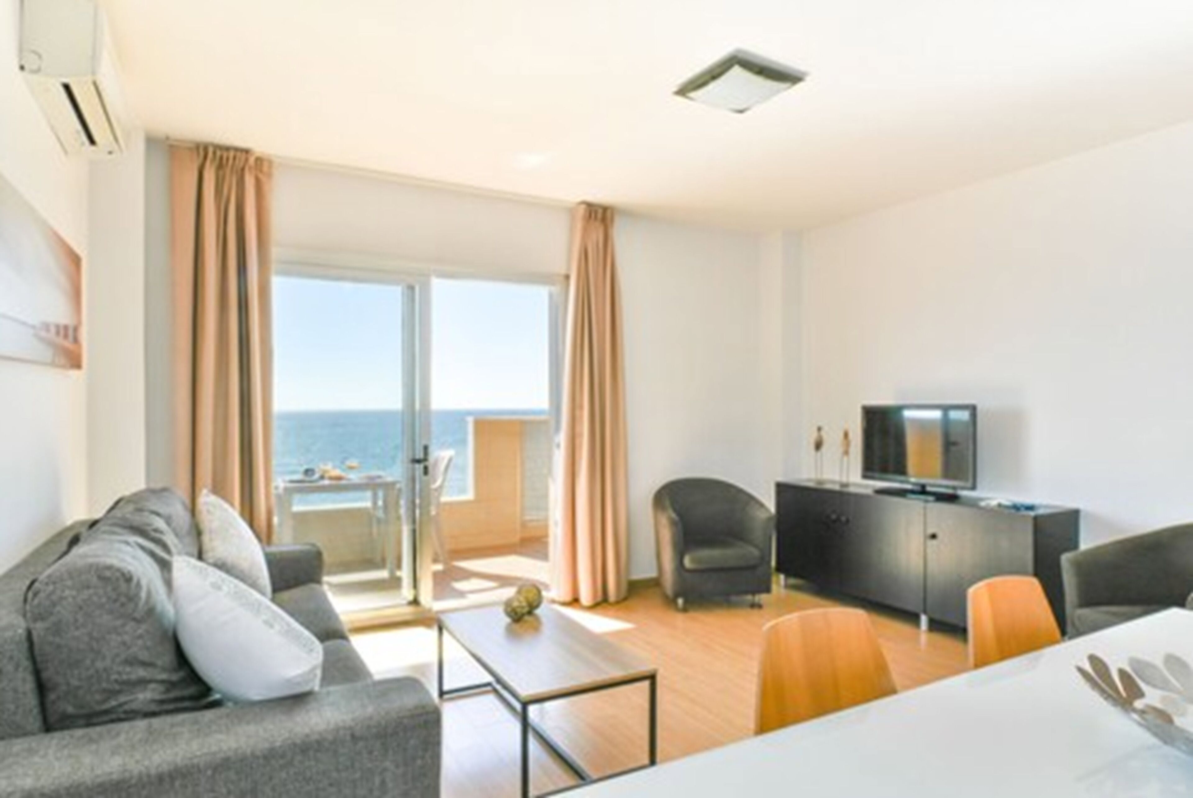 A modern living room with a sofa, a dining table, and a TV. There is a balcony with a view of the sea.