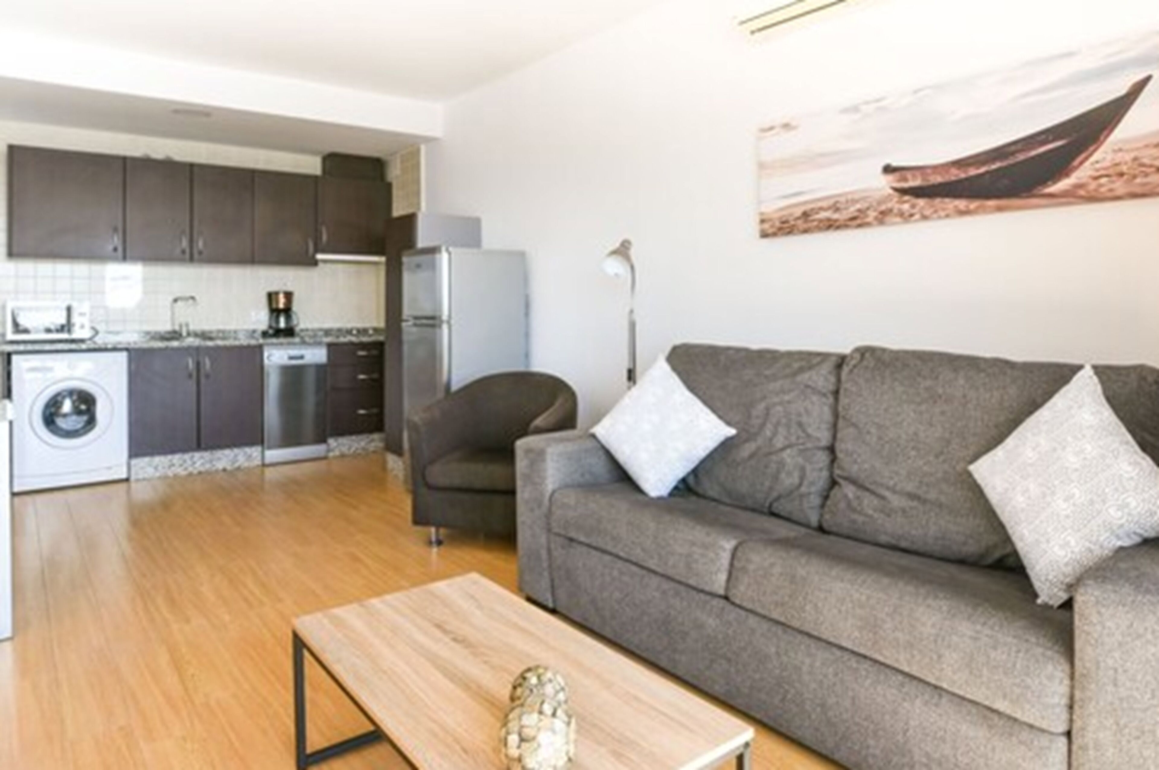 Un apartamento moderno con un sofá gris, una mesa de centro de madera, una cocina con armarios oscuros, una lavadora y un cuadro de un barco en la pared.