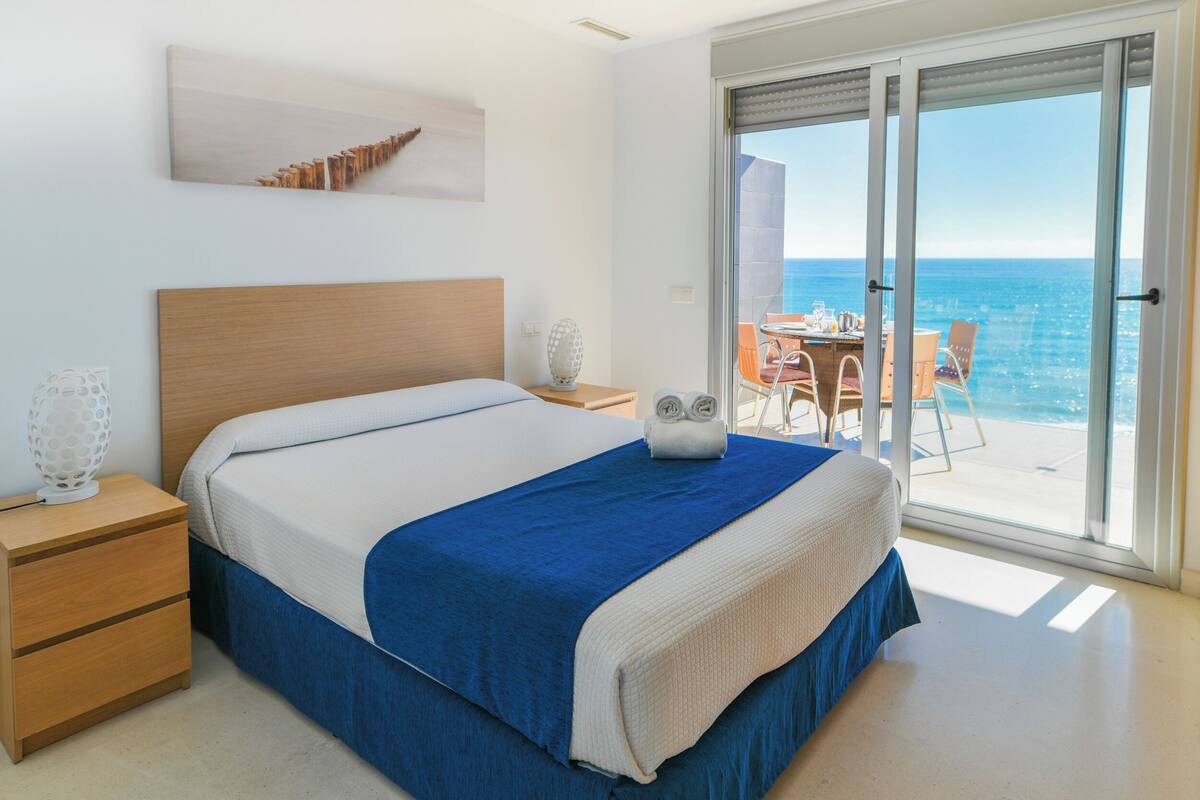 A hotel room with a bed, a nightstand, a table with chairs, and a view of the ocean through large sliding doors.