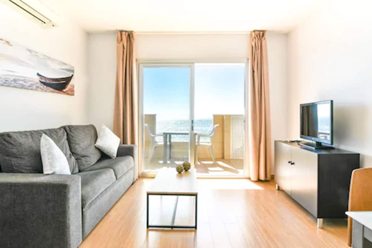 A modern living room with a grey sofa, a small coffee table, a TV on a cabinet, and a balcony with a view.