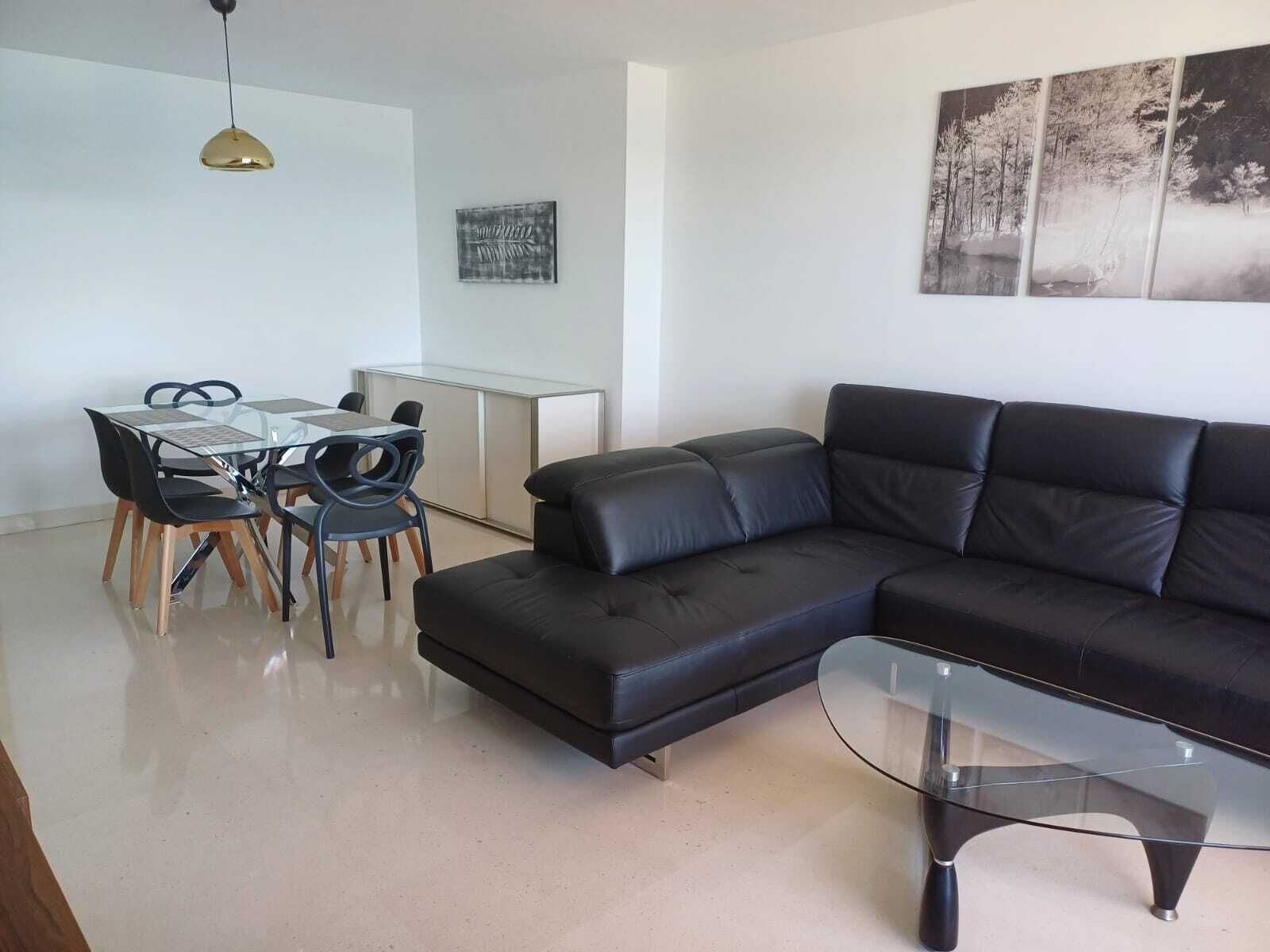 A modern living room with a black leather sofa, a glass coffee table, and a dining area with a glass table and chairs.