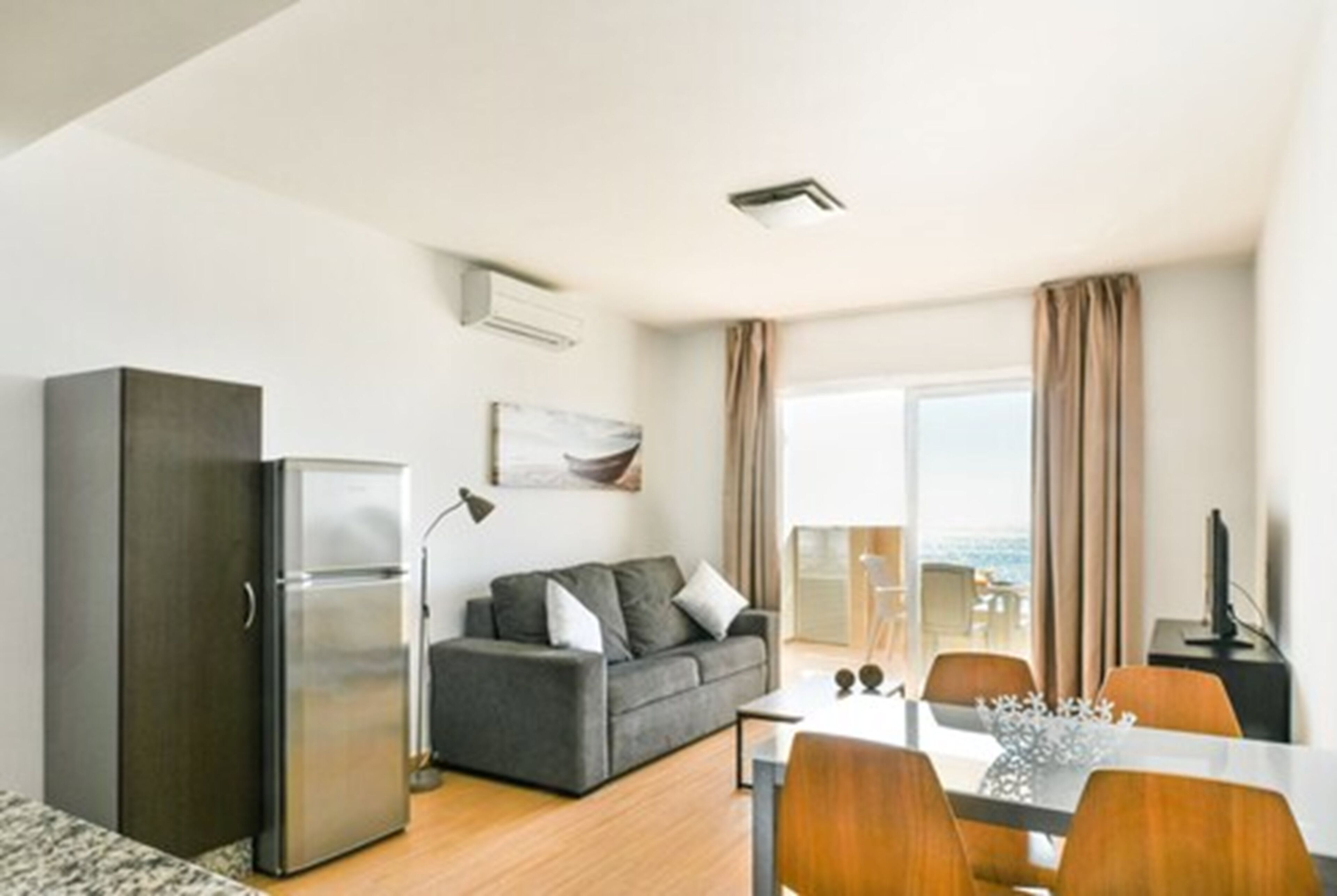 Een moderne woonkamer met een grijze bank, een eettafel met stoelen, een koelkast en een flatscreen-televisie.