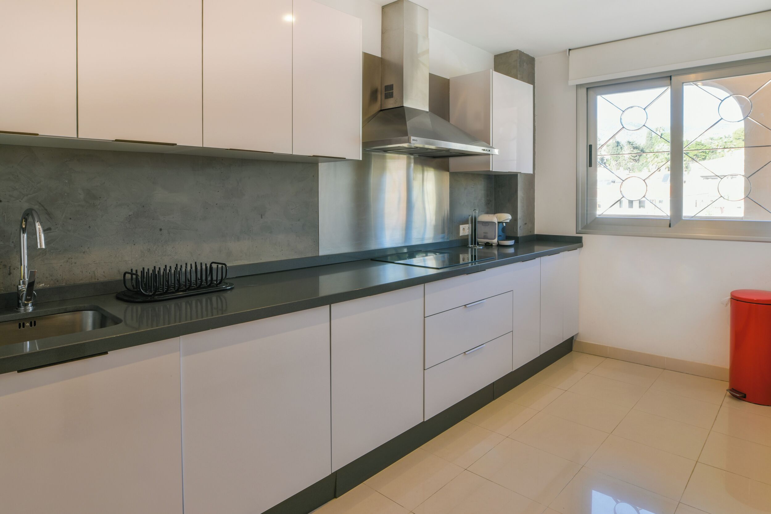 A modern kitchen with white cabinets, a stainless steel range hood, a built-in sink, and a window with a geometric design.