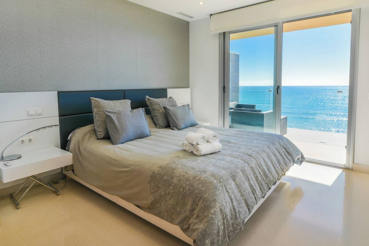 A modern bedroom with a large bed, a bedside table, and a view of the ocean through a sliding glass door.