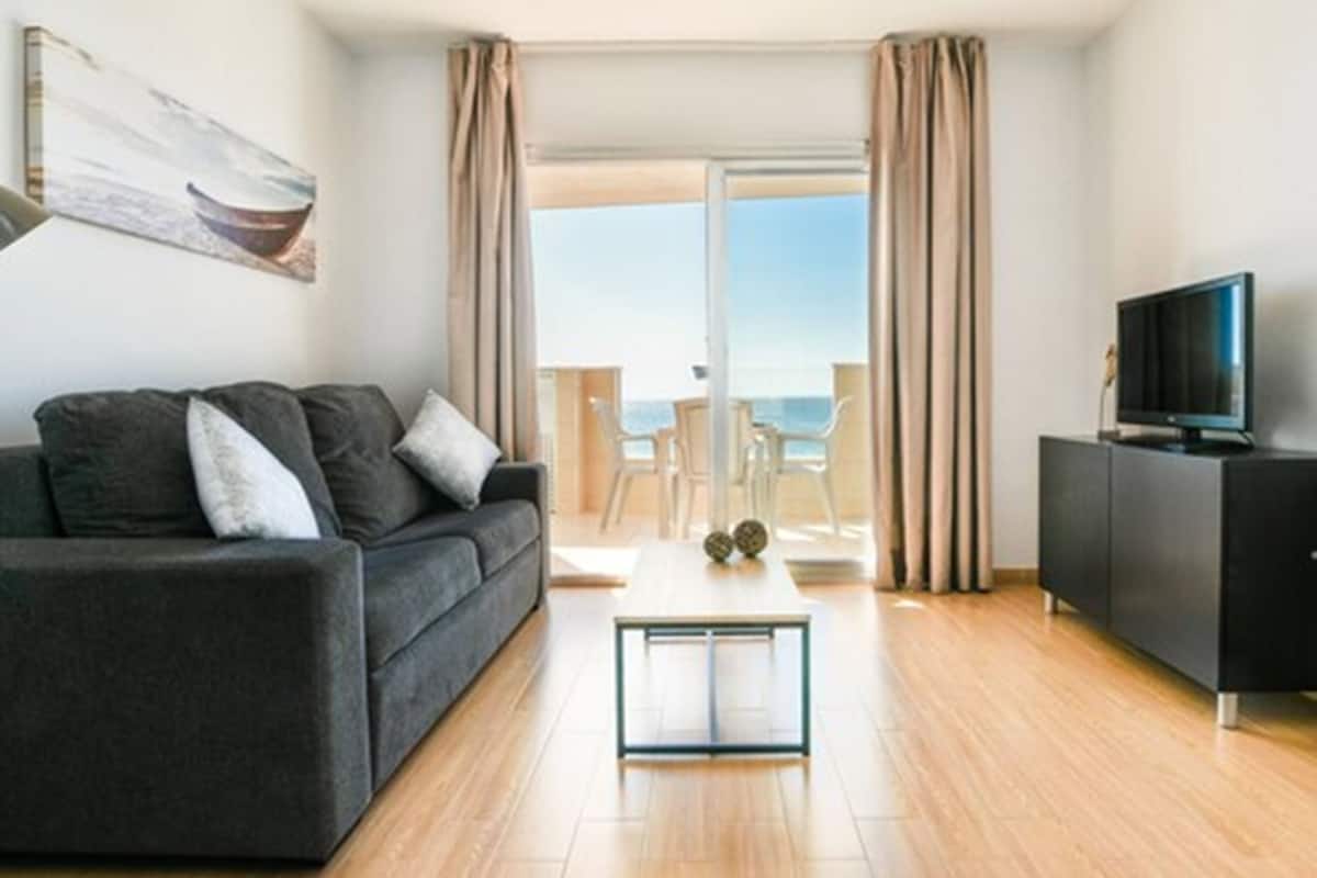 A modern living room with a sofa, a coffee table, a TV, and a balcony with a view of the sea.