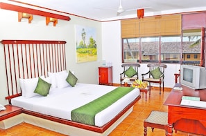 A hotel room with a large bed, two chairs, a small table, a television, and a window with a view of buildings. - Club Koggala Village (Koggala)