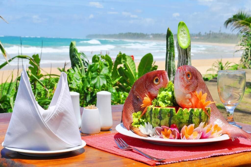 Prato de frutos do mar com dois peixes sobre um prato, cercado por legumes e guarnições, sobre uma mesa com guardanapo vermelho e saleiros, com vista para uma praia, oceano e vegetação.