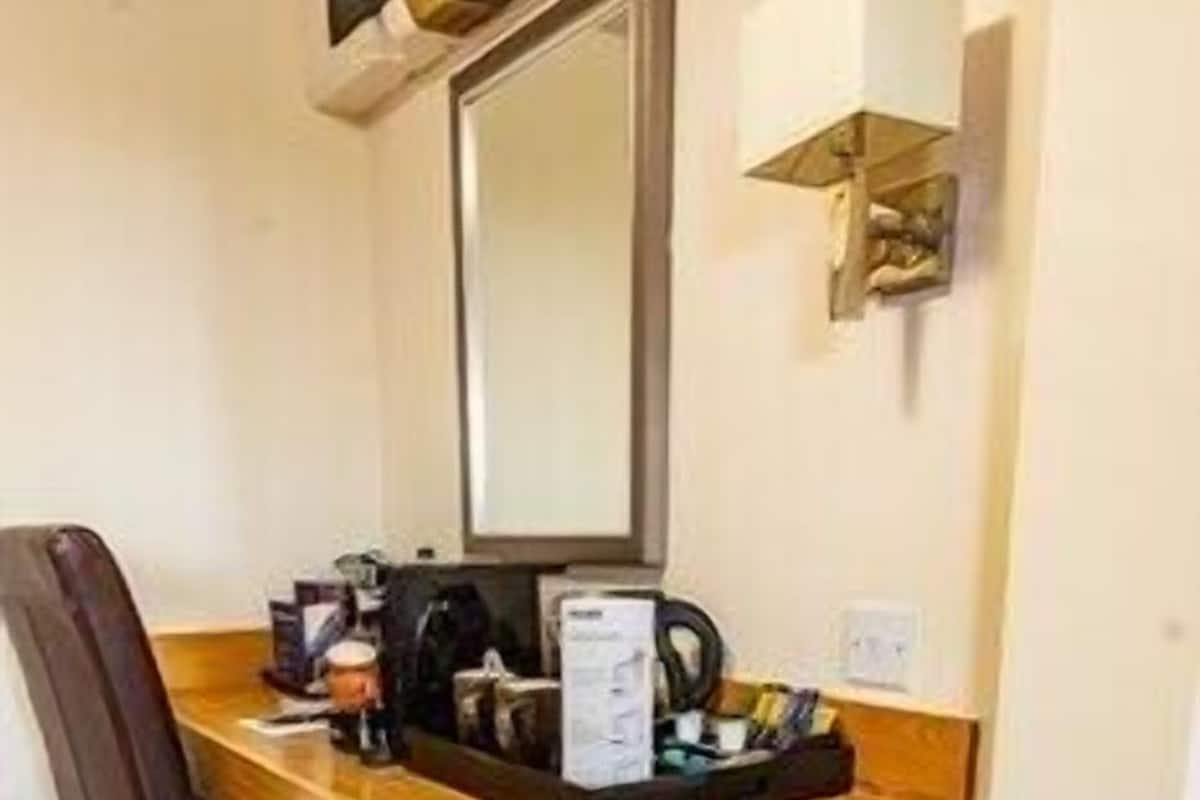 Une chambre d’hôtel de taille réduite, dotée d’un bureau en bois, d’une chaise, d’une petite poubelle et d’une lampe fixée au mur.