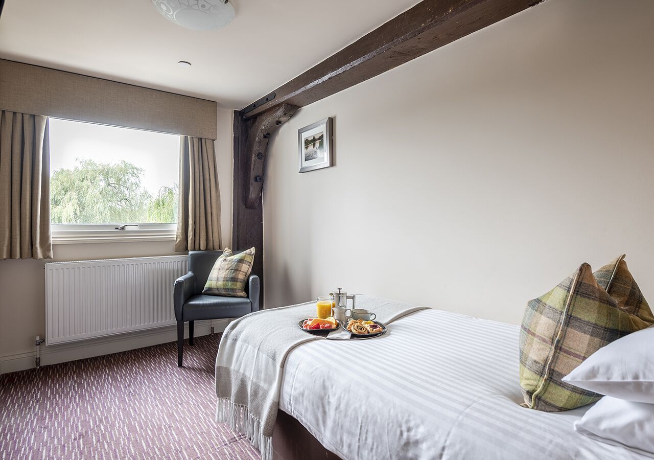 A hotel room with a bed, a chair, a window with curtains, a radiator, and a breakfast tray on the bed.