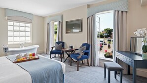 A hotel room with a bed, a desk, two chairs, a small table, a TV, a window with curtains, and a bathtub. - The Mill Hotel (Sudbury)