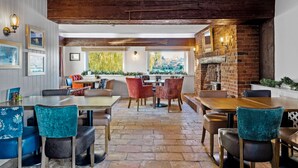 A restaurant with wooden beams, brick walls, and a stone floor. There are tables and chairs with blue and brown upholstery. - The Mill Hotel (Sudbury)