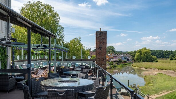 An outdoor patio with wicker furniture, a glass table, and a view of a river and greenery. - The Mill Hotel (Sudbury)