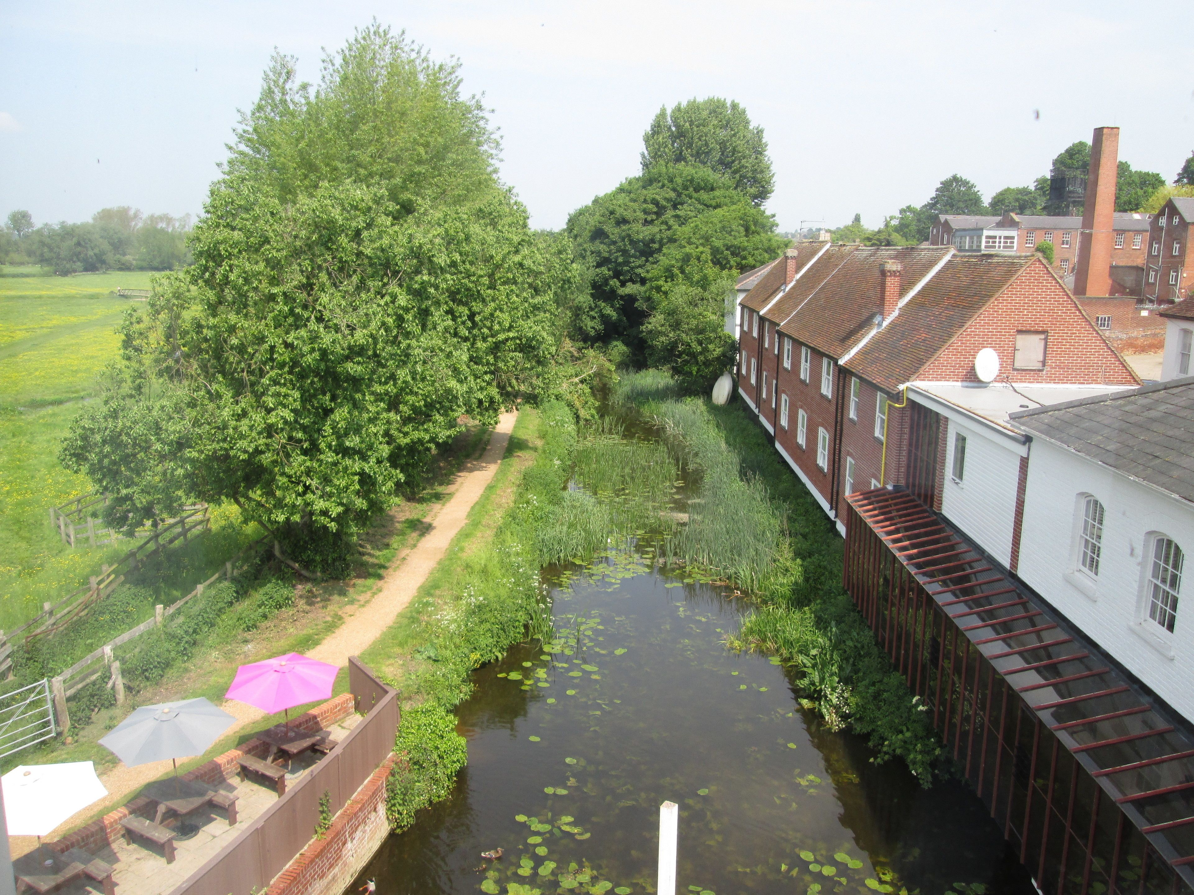 A narrow canal with a wooden walkway, surrounded by lush greenery and traditional brick buildings.