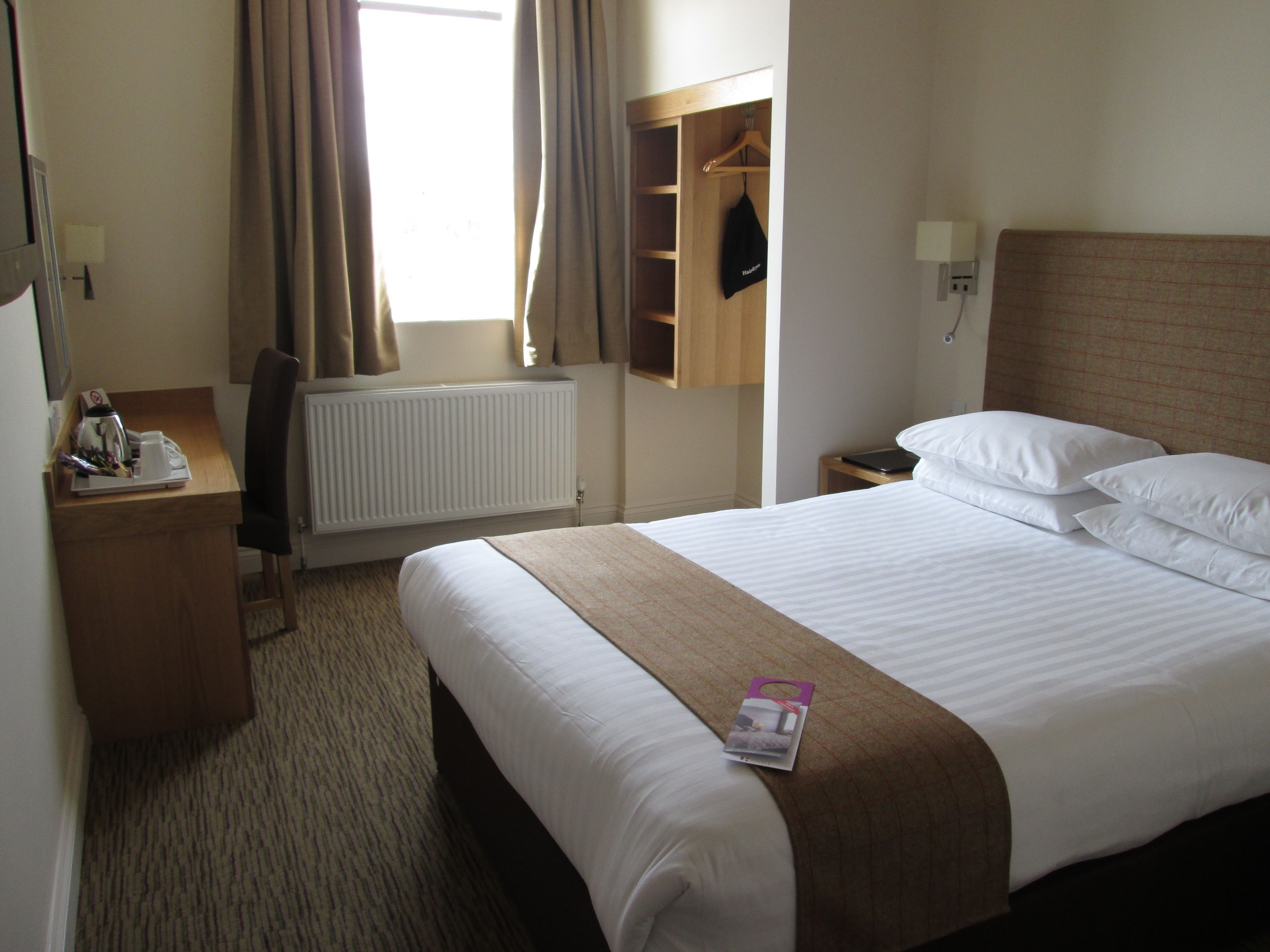 A hotel room with a bed, a bedside table, a lamp, a radiator, a window with curtains, and a coat hanging on a rack.
