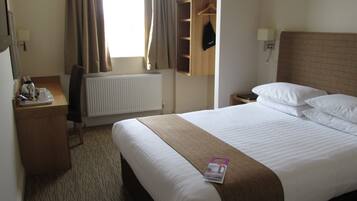 A hotel room with a bed, a bedside table, a lamp, a radiator, a window with curtains, and a coat hanging on a rack.