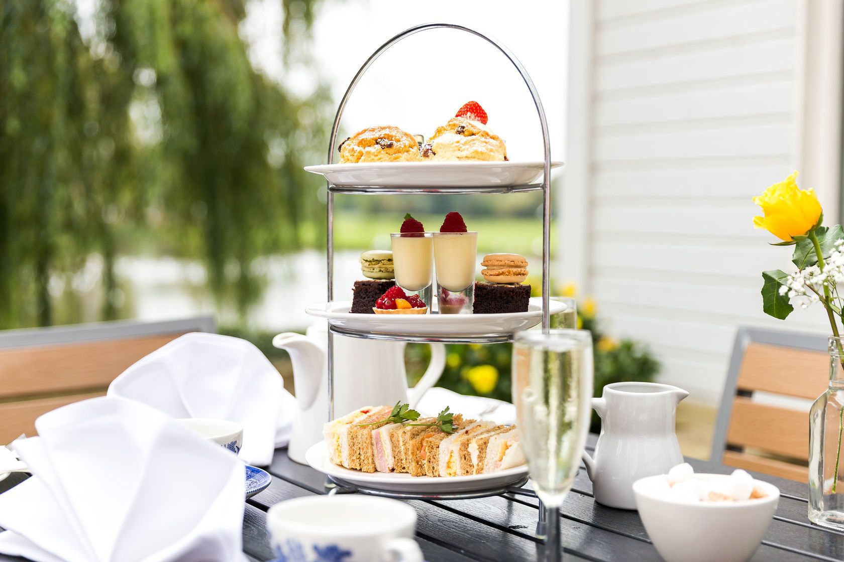A three-tiered dessert stand with various pastries and desserts, a teapot, cups, and a vase with a yellow flower on a wooden table outdoors.
