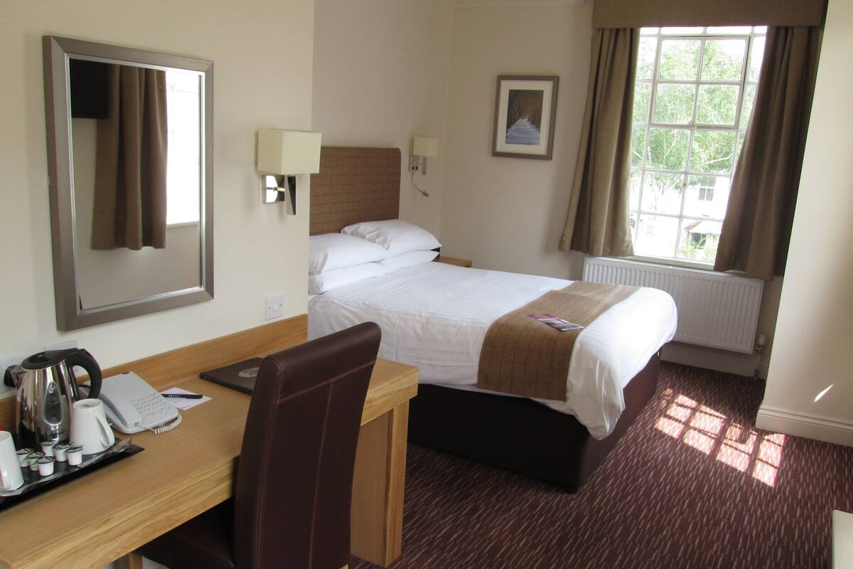 Une chambre d’hôtel comprenant un lit, un bureau avec une chaise, un miroir, une fenêtre avec des rideaux et un radiateur.